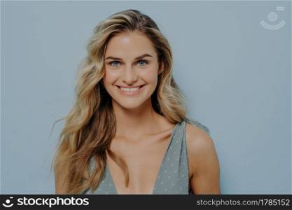 Happy youthful blonde girl in blue dress smiling with bright and cheerful smile, showing teeth, looking confident and positive while standing isolated over blue background in studio. Happiness concept. Happy youthful blonde girl smiling with bright and cheerful smile