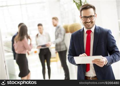 Hardworking businessman dressed in suit standing in modern office and using tablet while other businesspeople talking in background