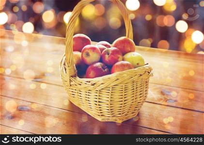harvest, season, autumn and fruits concept - close up of wicker basket with ripe red apples and leaves on wooden table over holidays lights
