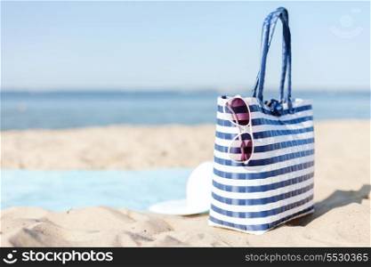 hats and summer concept - white straw hat, sunglasses and bag lying in the sand on the beach