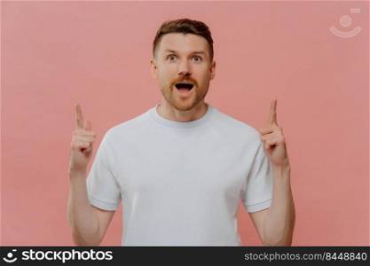 Having idea. Amazed excited young unshaven guy in white tshirt pointing with two forefingers up at empty copy space while getting answer on question or find out solution, isolated over pink background. Excited young man pointing with two forefingers up and with shocked face expression