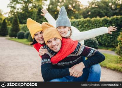 Having nice time together  Smiling excited woman, man and their little female child, wear warm knitted clothes, embrace each other, walk in park, being in good mood. Family give support, encouragement