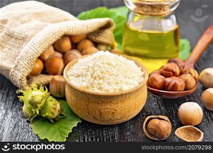 Hazelnut flour in a bowl, nuts in bag, a spoon, oil in glass jar and filbert branch with green leaves on the background of dark wooden board