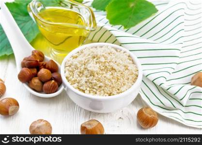 Hazelnut flour in a bowl, oil in a glass gravy boat, nuts, napkin and filbert branch with leaves on wooden board background