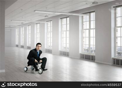 He is acting like a child. Young funny businessman riding three wheeled bicycle in office