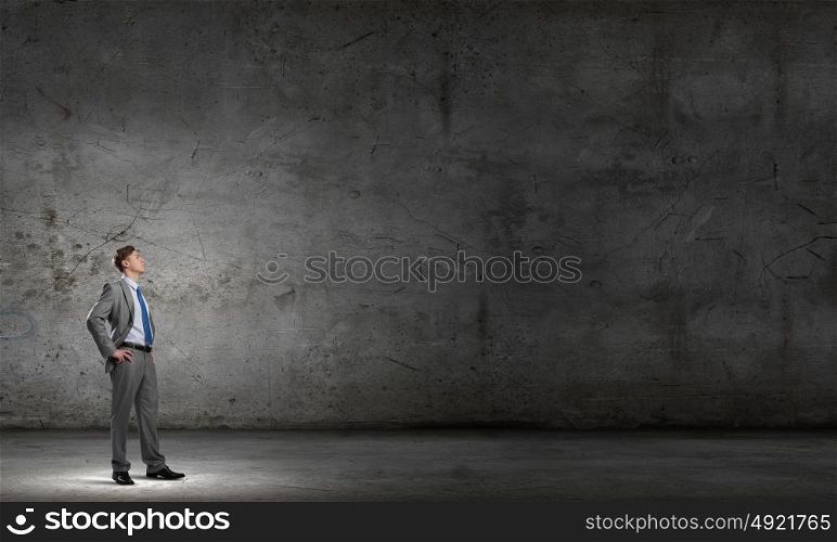 He is confident in his success. Confident young businessman with arms on waist in concrete room