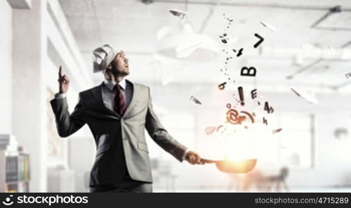 He is cooking something special. Young man in business suit and cook hat holding pan