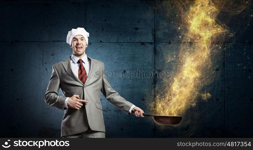 He is cooking something special. Young man in business suit and cook hat holding pan