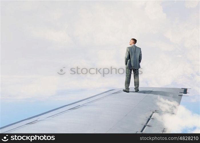 He is flying high. Young businessman standing on edge of airplane wing