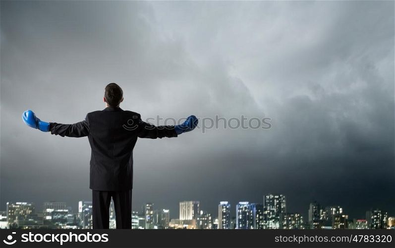 He is ready to fight for success. Back view of businessman in blue boxing gloves on city background