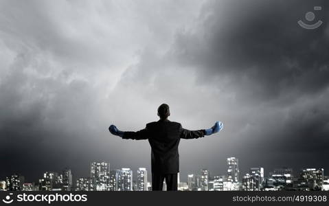 He is ready to fight for success. Back view of businessman in blue boxing gloves on city background