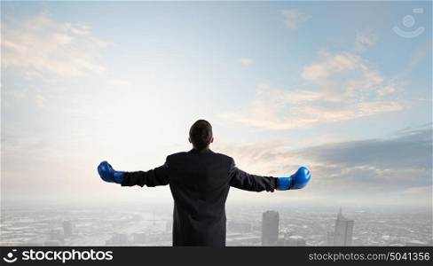 He is ready to fight for success. Back view of businessman in blue boxing gloves on city background