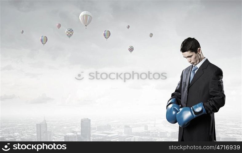 He is ready to fight for success. Young businessman in blue boxing gloves on city background