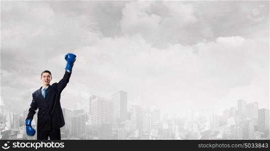 He is ready to fight for success. Young businessman in blue boxing gloves on city background