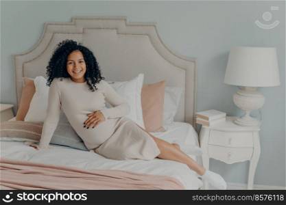 Hea<hy p®nant african american woman with curly hair relaxing on bed at home whi≤being on third trimester, dressed in white dress looking hapπly at camera and≥tting ready for motheρod. Happy p®nant african american lady resting on bed in modern apartment