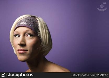 Head and shoulder portrait of bare young adult Caucasian blond woman on purple background wearing headband.