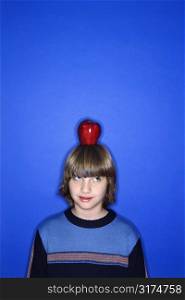Head and shoulder portrait of Caucasian boy with apple on his head standing against blue background.