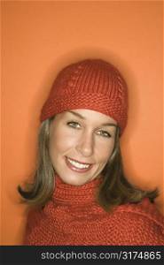 Head and shoulder portrait of smiling young adult Caucasian woman on orange background wearing winter hat and scarf.