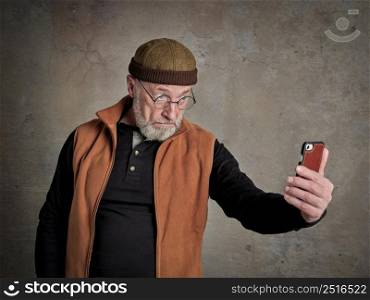 Head and shoulders portrait of a senior, bearded man taking selfie with his cell phone and making funny faces