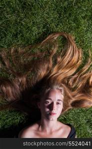 Head and shoulders shot of the beautiful young girl is lying on green grass in the evening time. Looking at camera. Blank expression on the face