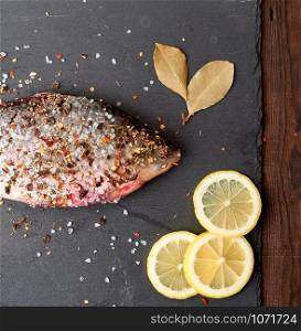 head of fresh crucian fish sprinkled with spices lies on a black slate board, top view