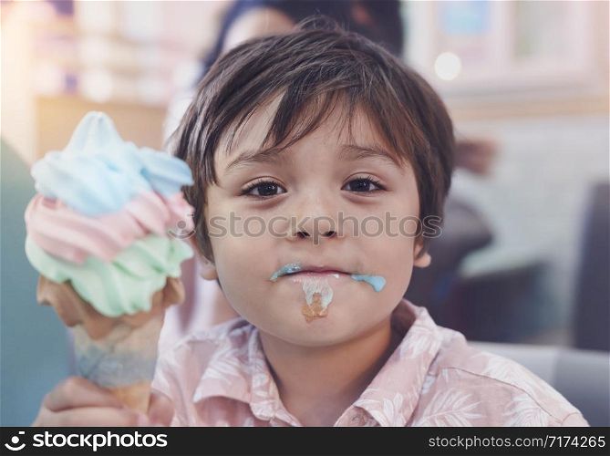 Head shot happy kid boy eating ice cream in the shop. Cute toddler with smiling face enjoy eating colourful ice cream on waffle cone. Candid shot children having fun in cafe