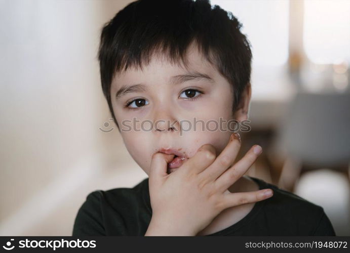 Head shot happy young kid looking at camera while sucking thumb after eating chocolate cake, Close up face cute child boy licking dirty fingers from eating sweet chocolate with blurry light background