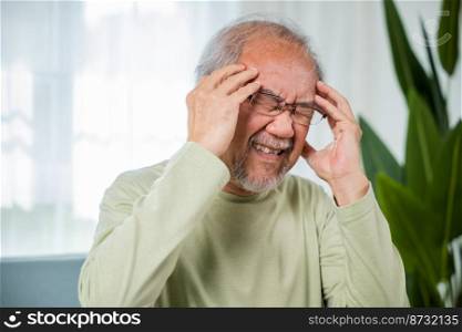 Headache. Close up of elderly holds head with hand suffering from migraine headache, Sad Asian senior man sitting on sofa feeling hurt and lonely at home, Old age health problems, healthcare