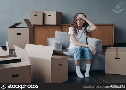 Headache from moving. Exhausted woman sitting among packed cardboard boxes. Spanish young lady in jeans and white t-shirt going to relocate. Depression and stress from moving concept.. Headache from moving. Exhausted spanish woman sitting among packed cardboard boxes.