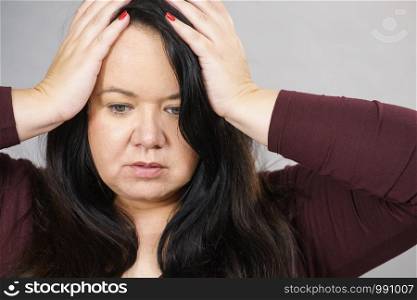 Headache, migraine and stress. Worried plus size adult upset woman suffering from head pain, studio shot on gray.. Worried woman having painfull headache