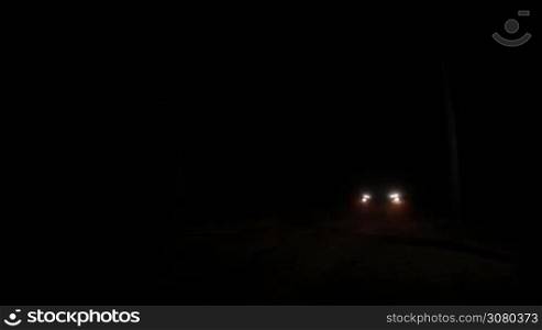 Headlights of car approaching from the dark. Car driving through the forest with yellow headlights illuminating rural dirt road at night. Night scene on the road in the woods.