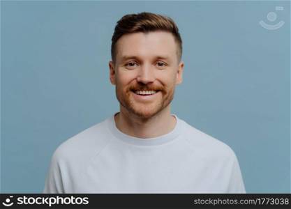 Headpshot portrait of young happy unshaven man in white tshirt looking at camera with excited facial expresson while posing against blue studio background. Human emotions and positivity concept. Young happy handsome unshaven man in casual wear smiling at camera