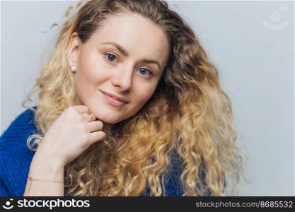Headshot of adorable blue eyed female model with curly hair, blue eyes, holds chin, looks with pleased expression as listens interlocutor with great interest, isolated over blue studio background