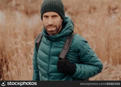 Headshot of atrractive blue eyed man has dark stubble, wears hat, jacket, gloves, carries rucksack, has outdoor stroll, models against blurred background. People, leisure, traveling and rest concept
