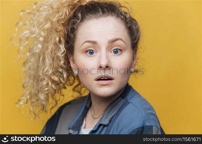 Headshot of attractive blue eyed woman in bewilderment, has stunned expression, can`t understand what she should do, looks with puzzlement directly into camera, isolated over yellow background