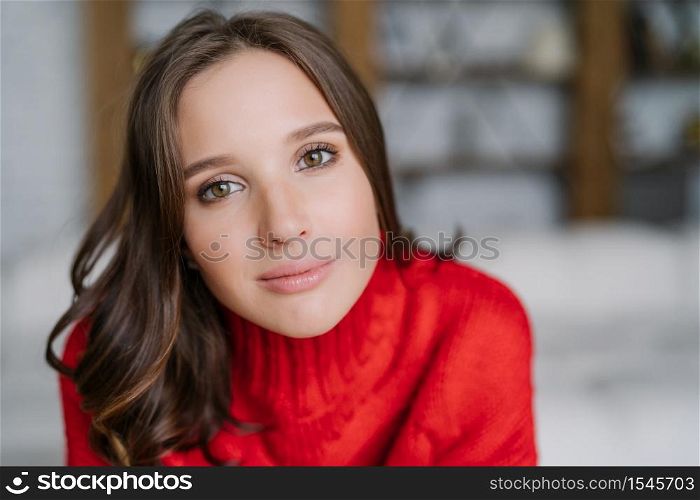Headshot of attractive healthy European woman with dark hair and soft skin, looks directly at camera, wears warm red sweater, models indoor. People and lifestyle concept. Domestic atmosphere