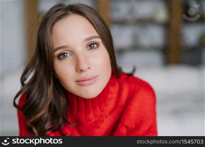 Headshot of attractive healthy European woman with dark hair and soft skin, looks directly at camera, wears warm red sweater, models indoor. People and lifestyle concept. Domestic atmosphere
