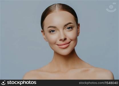 Headshot of attractive woman has beautiful facial features, natural makeup, dark combed hair, naked body, full lips, isolated on blue background, undergoes beauty treatments. Skin care concept