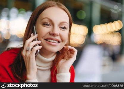Headshot of attractive young woman with glad facial expression, talks via cell phone, has brown hair, happy to hear news, has free time, blurred background with copy space for your promotion