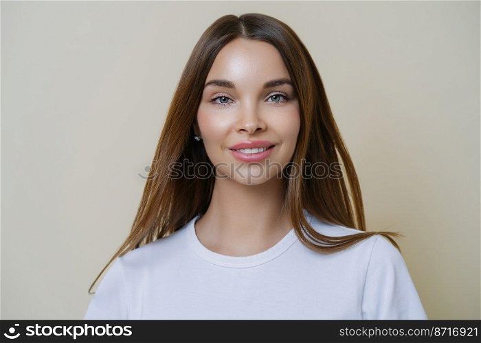 Headshot of beautiful dark haired young European woman looks with tender expression, has friendly attitude, talks casually with friends, wears casual white t shirt, isolated over beige background