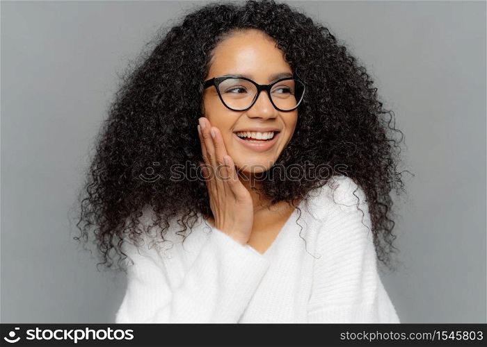 Headshot of beautiful dark skinned female model touches cheek gently, has charming smile on face, looks happily away, has curly Afro hair, wears spectacles and white jumper, isolated on grey wall