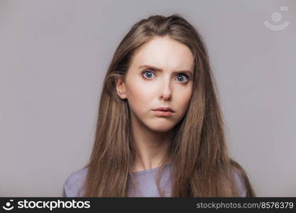 Headshot of beautiful female with blue eyes and pure skin, looks seriously and angrily at camera, has dark straight hair, isolated over grey background. Horizontal shot of lovely young woman indoor