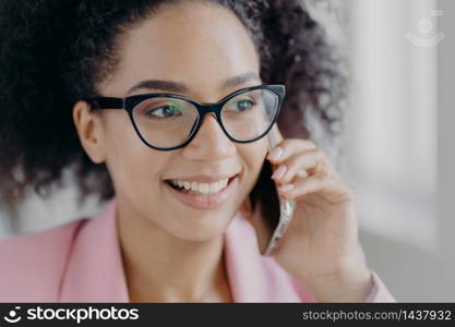 Headshot of beautiful smiling dark skinned woman wears optical glasses, holds modern cell phone, has well cared complexion, talks about future investments with business partner, looks aside.