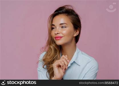 Headshot of beautiful young woman has minimal makeup, looks refreshed, turns aside, has satisfied thoughtful expression, wears blue shirt, thinks about weekend, isolated against purple background.