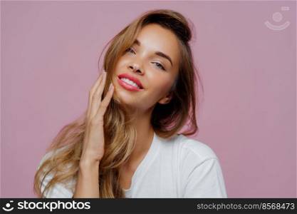 Headshot of carefree relaxed feminine lady tilts head left, touches her long wavy hair, wears lipstick and makeup, prepares for special event in her life, being professional photo model, stands indoor