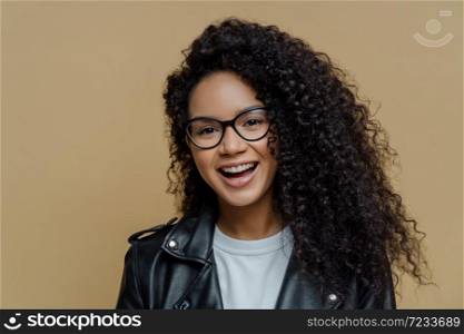 Headshot of cheerful African American woman with dark curly hair, has pleased face expression, joyful conversation, wears transparent glasses and black leather jacket, isolated on beige background