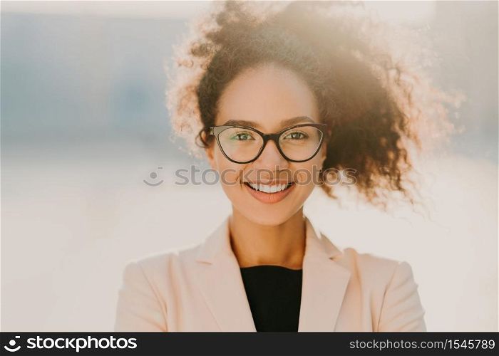 Headshot of cheerful curly woman with positive expression, wears spectacles, white elegant jacket, looks straightly at camera, stands outdoor, has curly hair expresses good emotions. Ethnicity concept