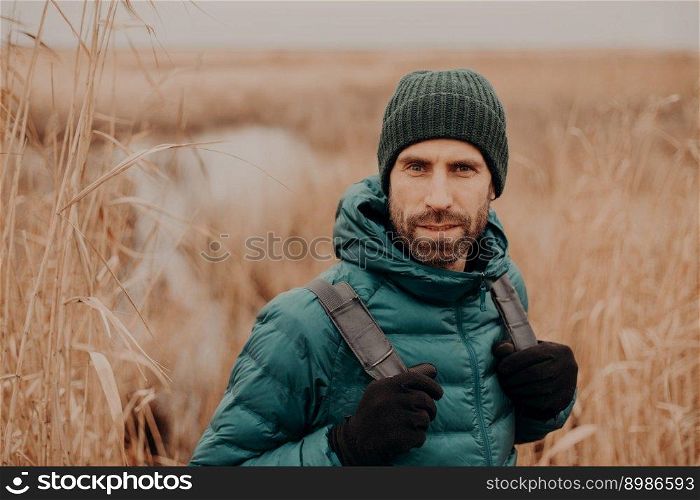 Headshot of confident unshaven guy in hat and jacket, carries rucksack, poses against autumn nature background, has outdoor stroll.