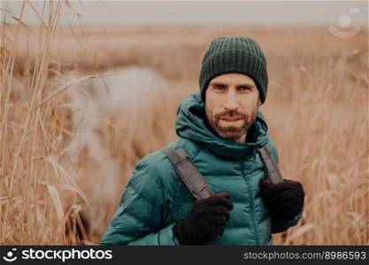 Headshot of confident unshaven guy in hat and jacket, carries rucksack, poses against autumn nature background, has outdoor stroll.
