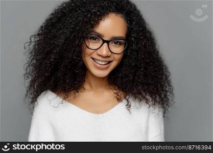 Headshot of curly haired smiling woman has healthy dark skin, Afro hairstyle, smiles gently at camera, weras optical glasses and white jumper enjoys spare time with friends isolated on grey background